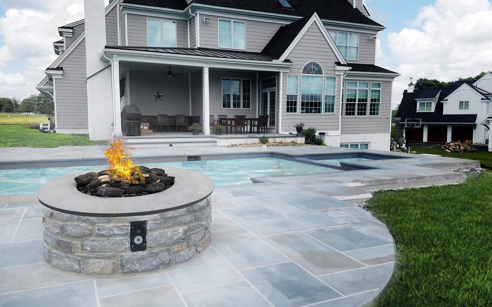 A propane fire pit burns on a flagstone patio next to a backyard pool.